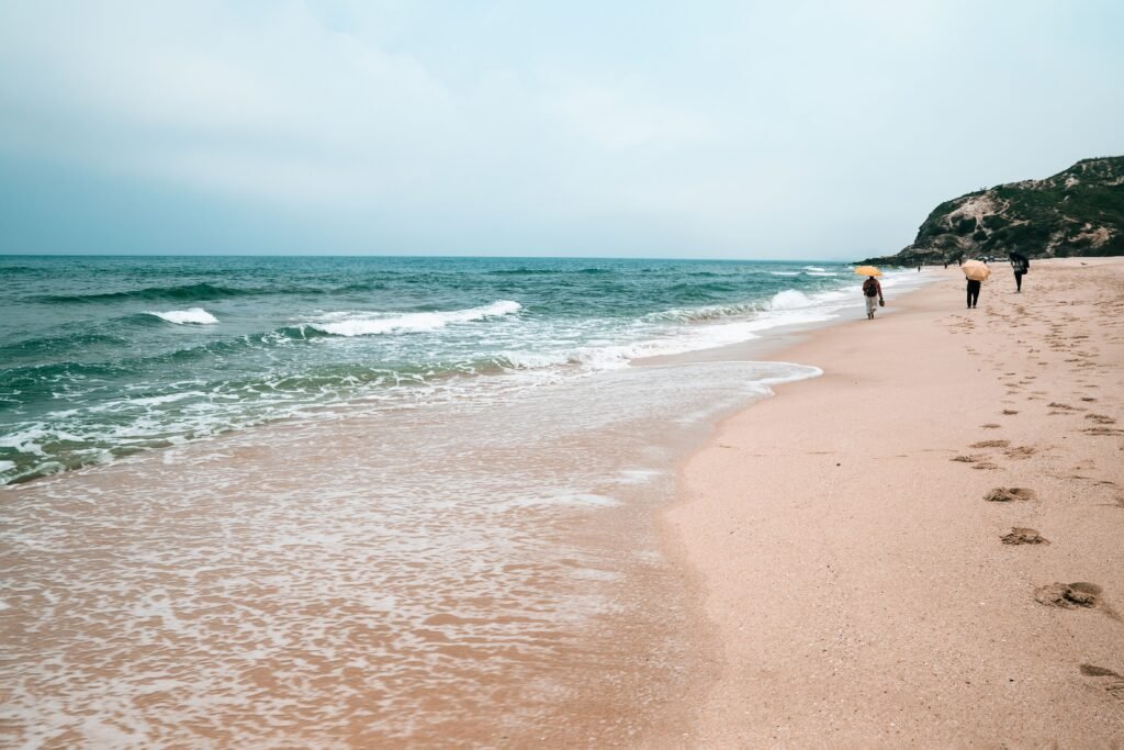 A tranquil beach with gentle waves lapping at the shore as people stroll along the sandy coastline under an overcast sky.